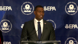 EPA administrator Michael Regan strands behind a translucent podium, wearing a black suit jacket, white collar shirt, navy blue tie and an American flag pin. The Navy blue background behind him is interspersed with white EPA logos. 